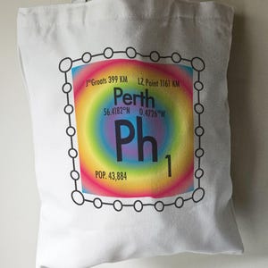 May include: A white tote bag featuring a black and white graphic of a periodic table element. The element is labelled "Ph" for Perth, with the city's coordinates and population listed. The background of the element is a rainbow gradient.