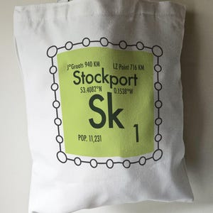 May include: A white tote bag featuring a green and black design. The design is a stylised periodic table element for Stockport, England. It includes the city's name, coordinates, population, and the element symbol "Sk".
