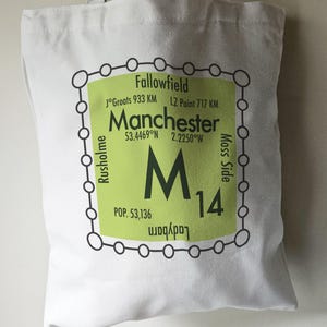 May include: A white tote bag with a green and black graphic design. The graphic features a stylized map of Manchester, England, with the city's name, coordinates, and population. The graphic also includes the letter "M" and the number "14".
