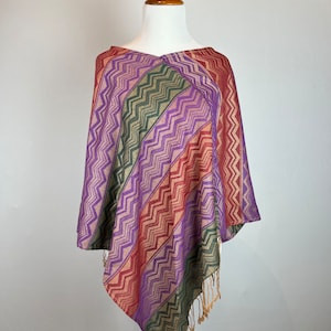 Op de afbeelding: Een poncho met een paars, groen en rood patroon en een franje rand. De poncho is gemaakt van een zachte, lichtgewicht stof en is perfect om te laagjes.