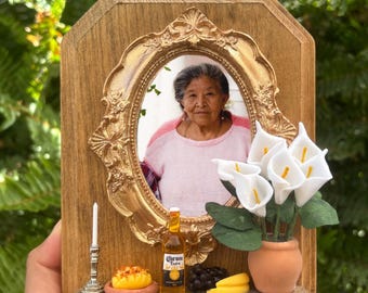 May include: A wooden memorial frame with a gold oval frame holding a photo of a woman. Miniature items include a candle, a bottle of Corona Extra, a small pot of white calla lilies, and colourful papel picado banners.