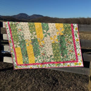 May include: A quilt with a patchwork design featuring yellow, green, and pink floral fabric. The quilt is hanging on a wooden fence against a backdrop of hills and a blue sky.