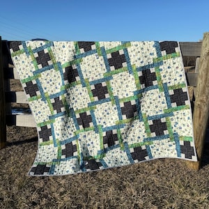 Może przedstawiać: Kołdra z geometrycznym wzorem w kolorze czarnym, zielonym i białym. Kołdra jest wykonana z tkaniny z kwiatowym nadrukiem. Kołdra jest zawieszona na drewnianym płocie.