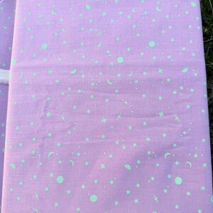 Pode incluir: Tecido dobrado com fundo lavanda e um padrão repetido de estrelas, luas e círculos verde claro. O tecido é provavelmente algodão ou material semelhante, adequado para projetos de costura ou artesanato.