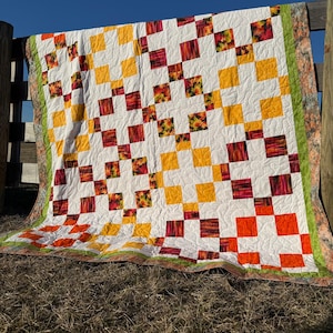 Könnte beinhalten: Eine Steppdecke mit Patchwork-Muster aus Quadraten in Weiß, Gelb, Orange und Rot mit floralem Muster. Die Steppdecke hängt an einem Holzzaun.