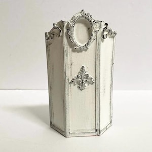 May include: A white, hexagonal decorative container with ornate silver accents. Features an oval frame and a fleur-de-lis design. The container has a distressed, vintage appearance, with dark gray trim.