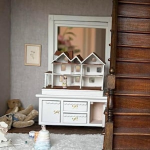 May include: A miniature dollhouse with a white facade and brown roof sits atop a white cabinet with gold bow accents. A wooden staircase and a teddy bear are also visible. The scene is set in a room with a light fixture.