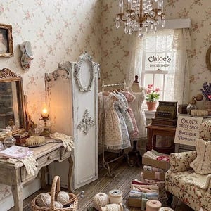 May include: A miniature dollhouse room depicting Chloe's Dress Shop with floral wallpaper, a white vanity, a tall armoire, a clothing rack with small dresses, a mannequin, a cash register, and a sign reading Chloe's Dress Shop Dresses & Trimmings.