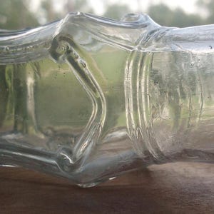 May include: A clear glass bottle with a rectangular body and a rounded neck. The bottle has a textured surface and a wide opening. The bottle is lying on a wooden surface.