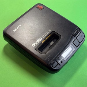 Pu&ograve; includere: Un lettore CD Sony Discman nero con un logo 'Discman' argentato e un display digitale. Il lettore ha un pulsante etichettato 'Open' e un pulsante etichettato 'Play Mode'.