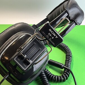 Pu&ograve; includere: Cuffie Sony DR-9 nere con cavo a spirale e manopola di controllo del volume e del tono. Le cuffie hanno un padiglione in pelle nera e un'archetto in metallo argentato. L'etichetta sulle cuffie recita "SONY DR-9 IMP 40 CON MADE IN JAPAN".