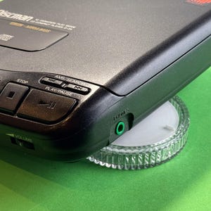 Pu&ograve; includere: Un lettore CD portatile Discman nero con una placca frontale e pulsanti argentati. Il lettore ha un display digitale, una manopola del volume e una presa per le cuffie. Il lettore &egrave; posizionato su un supporto in plastica trasparente.