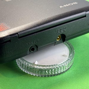 Pu&ograve; includere: Un lettore cassette Sony nero con un jack di uscita linea e un jack di ingresso DC 6V. Il lettore &egrave; posizionato su un supporto in plastica trasparente.