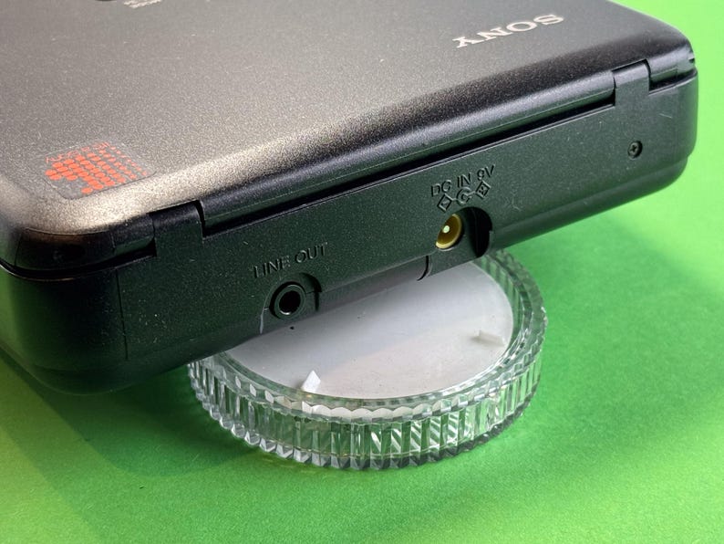 Pu&ograve; includere: Un lettore cassette Sony nero con un jack di uscita linea e un jack di ingresso DC 6V. Il lettore &egrave; posizionato su un supporto in plastica trasparente.