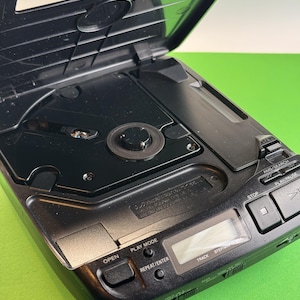Pu&ograve; includere: Lettore CD portatile nero con uno schermo di visualizzazione argentato e pulsanti etichettati "OPEN", "PLAY MODE", "REPEAT/ENTER", "TRACK", "STEP", "STOP", "AMS/SEARCH" e "PLAY/PAUSE".