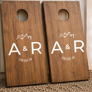 May include: Two rectangular wooden cornhole boards with a dark brown wood grain finish. Each board features a circular hole at the top and a white graphic decal displaying the letters A & R, a small leafy branch icon, and the date 08.08.26.