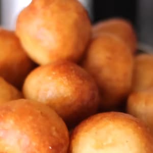 May include: A close-up shot of a pile of golden-brown, round pastries. The pastries appear to be freshly fried, with a slightly glossy surface. They are arranged in a metal strainer, suggesting they are ready to be served or cooled.