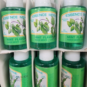 Puede incluir: Una exhibición de botellas verdes de Essence Menth con tapas blancas. Las etiquetas presentan ilustraciones de hojas de menta y texto multilingüe, incluyendo "Mint Flavor" y "Sin Alcohol".