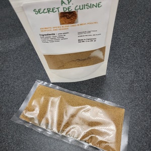 Könnte beinhalten: Ein weißer wiederverschließbarer Beutel mit einer braunen Gewürzmischung. Das Etikett lautet "A.P. Secret de Cuisine" und listet die Zutaten auf: schwarzer Pfeffer, Nelke, Kräuter der Provence, Paprika, Muskatnuss, Curry, Landzwiebeln, Jamaika-Muskatnuss und grüner Anis. Das Etikett gibt auch an "Aromatische Gewürzmischung für Fleisch, Geflügel, Meeresfrüchte und Gemüse" und "Hergestellt in Kamerun, Nettogewicht 2 oz (50g)".