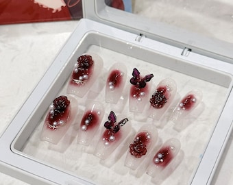 Può includere: Un set di unghie finte in una scatola bianca. Le unghie presentano una base trasparente con un design sfumato bordeaux, adornate con rose bordeaux, perle e decorazioni a farfalla viola. Le unghie sono di forma ovale.