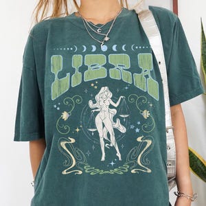 May include: Dark teal t-shirt with a green "LIBRA" graphic, a female figure with scales, and celestial designs. The tee has a vintage appearance. The design is printed on the front.