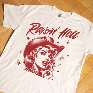 May include: Off-white t-shirt with a red graphic. The graphic depicts a woman in a cowboy hat smoking, with the words "Raisin' Hell" above. Small red stars are scattered around the image.