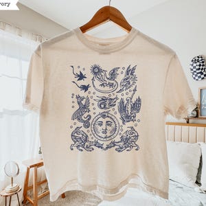 May include: An ivory short-sleeve t-shirt featuring a dark blue graphic print of various mythical dragons and a central sun face. The design includes multiple winged creatures arranged around a circular sun with a human-like facial expression.