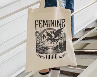 May include: A beige tote bag with a black and white graphic design of a dragon flying in front of a full moon. The text "FEMININE RAGE" is printed in black on the bag.