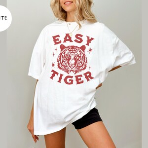 May include: White t-shirt with a red tiger graphic and the text "EASY TIGER". The shirt is a casual style, with short sleeves and a relaxed fit. The graphic features a detailed tiger face.
