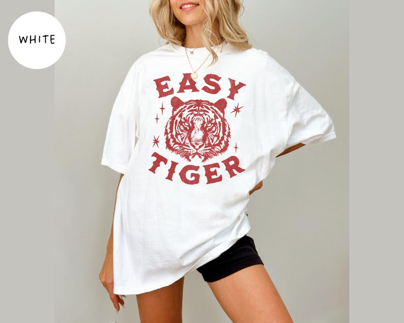 May include: White t-shirt with a red tiger graphic and the text "EASY TIGER". The shirt is a casual style, with short sleeves and a relaxed fit. The graphic features a detailed tiger face.
