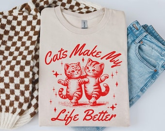 May include: Cream-colored t-shirt with the text "Cats Make My Life Better" in red script. Two red cartoon cats are embracing. The shirt is folded next to a brown and white checkered sweater and blue jeans.