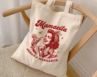 May include: Off-white canvas tote bag with red text and graphic. The text reads "Mamacita Needs a Margarita" above an illustration of a woman holding a cocktail. The bag has two handles and is sitting on a wooden chair.
