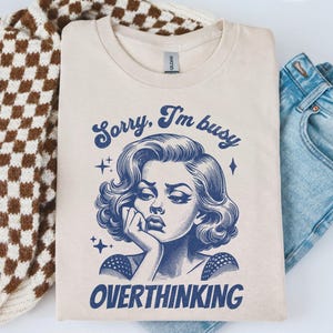May include: A sand-colored t-shirt with the text "Sorry, I'm busy OVERTHINKING" and a blue illustration of a woman. The shirt is folded next to a brown and white checkered sweater and blue jeans.