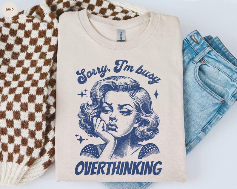 May include: A sand-colored t-shirt with the text "Sorry, I'm busy OVERTHINKING" and a blue illustration of a woman. The shirt is folded next to a brown and white checkered sweater and blue jeans.