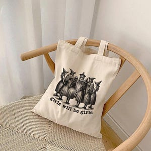 May include: A beige canvas tote bag with black handles and a black and white illustration of a group of witches with the text "girls will be girls".
