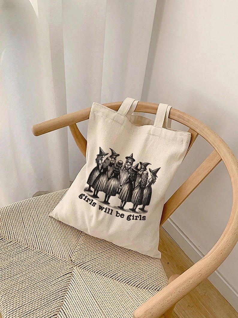 May include: A beige canvas tote bag with black handles and a black and white illustration of a group of witches with the text "girls will be girls".