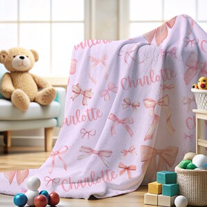 Pode incluir: Um cobertor de beb&ecirc; lil&aacute;s com um padr&atilde;o de la&ccedil;os em aquarela rosa e o nome "Charlotte" em escrita rosa. O cobertor est&aacute; sobre uma cadeira e uma mesa pequena, com um ursinho de pel&uacute;cia e brinquedos no fundo.