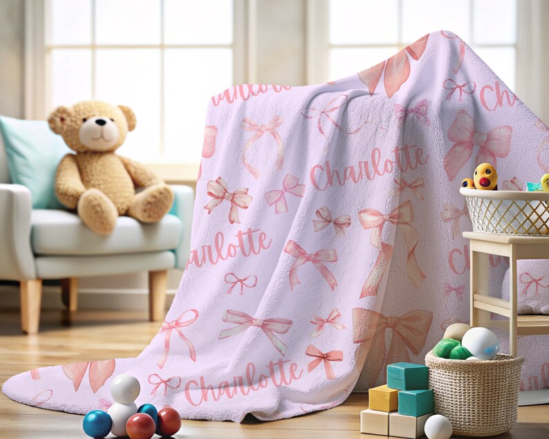 Pode incluir: Um cobertor de beb&ecirc; lil&aacute;s com um padr&atilde;o de la&ccedil;os em aquarela rosa e o nome "Charlotte" em escrita rosa. O cobertor est&aacute; sobre uma cadeira e uma mesa pequena, com um ursinho de pel&uacute;cia e brinquedos no fundo.