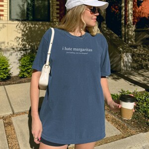 May include: A denim blue short-sleeve t-shirt featuring the white text "i hate margaritas" followed by the smaller text "just kidding, can you imagine?" centered on the chest. The shirt has a crew neckline and a relaxed, loose-fitting silhouette.