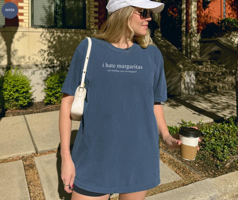 May include: A denim blue short-sleeve t-shirt featuring the white text "i hate margaritas" followed by the smaller text "just kidding, can you imagine?" centered on the chest. The shirt has a crew neckline and a relaxed, loose-fitting silhouette.