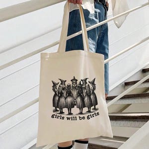 May include: A beige tote bag with a black and white illustration of a group of girls wearing witch hats and the text "girls will be girls".