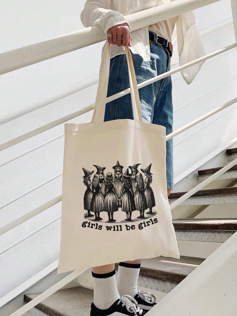 May include: A beige tote bag with a black and white illustration of a group of girls wearing witch hats and the text "girls will be girls".