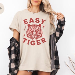 May include: A sand-colored t-shirt with the text "EASY TIGER" in red, featuring a tiger head graphic. The shirt is worn with a black and gray plaid shirt. The model is wearing a necklace.
