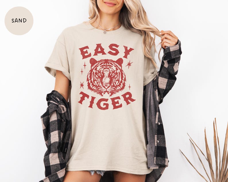 May include: A sand-colored t-shirt with the text "EASY TIGER" in red, featuring a tiger head graphic. The shirt is worn with a black and gray plaid shirt. The model is wearing a necklace.