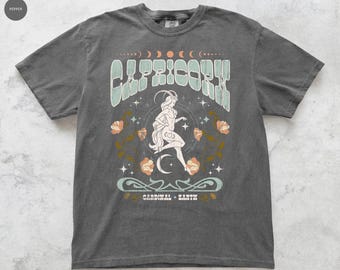 Könnte beinhalten: Dunkelgraues T-Shirt mit einem Steinbock-Design. Das Design zeigt das Wort "CAPRICORN" in hellgrüner Schrift, eine Frauenfigur, einen Halbmond, Sterne und florale Elemente in Orange und Hellgrün. Der Text "CARDINAL + EARTH" befindet sich unten.