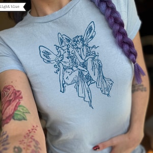 Puede incluir: Camiseta azul claro con una ilustración azul oscuro de dos hadas aladas. El texto "light blue" está en la esquina superior izquierda. La camiseta es de cuello redondo.