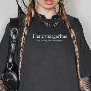May include: A black short-sleeved t-shirt featuring the white text "i hate margaritas" followed by the smaller text "just kidding, can you imagine?" centered on the chest. The shirt is worn with a silver chain necklace and a black bag.