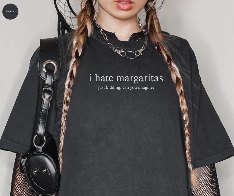 May include: A black short-sleeved t-shirt featuring the white text "i hate margaritas" followed by the smaller text "just kidding, can you imagine?" centered on the chest. The shirt is worn with a silver chain necklace and a black bag.