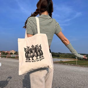 May include: A white canvas tote bag with a black and white illustration of a group of witches. The text "Girls will be girls" is printed on the bag.