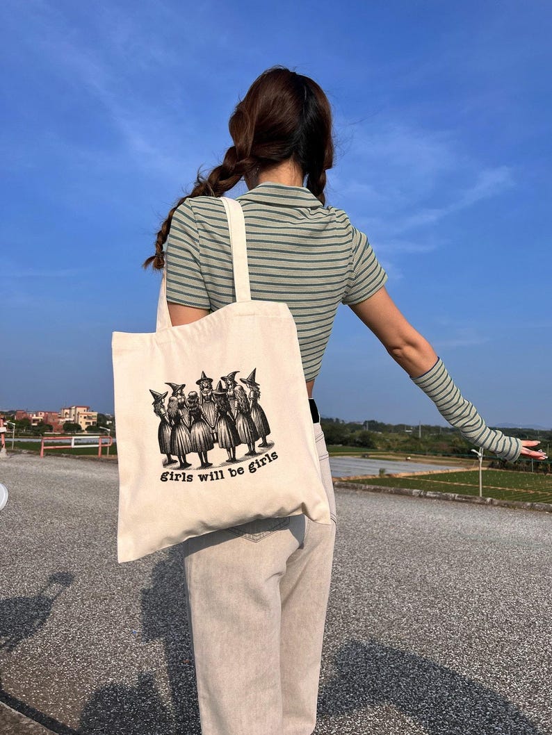 May include: A white canvas tote bag with a black and white illustration of a group of witches. The text "Girls will be girls" is printed on the bag.